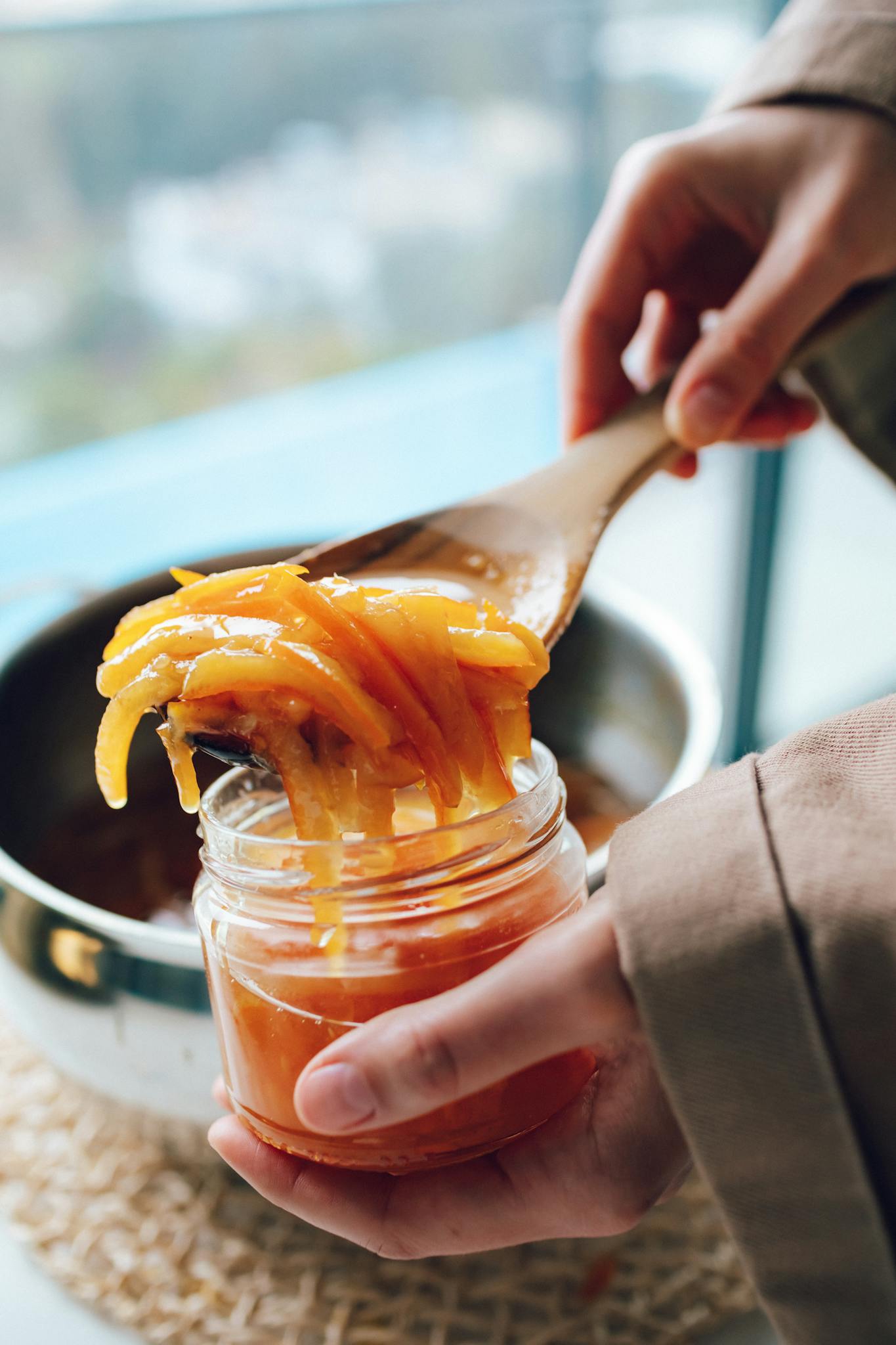 Hands preparing homemade orange marmalade with a wooden spoon and jar.