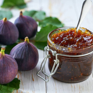 Glass jar of figs jam with fresh figs on a white wooden table.